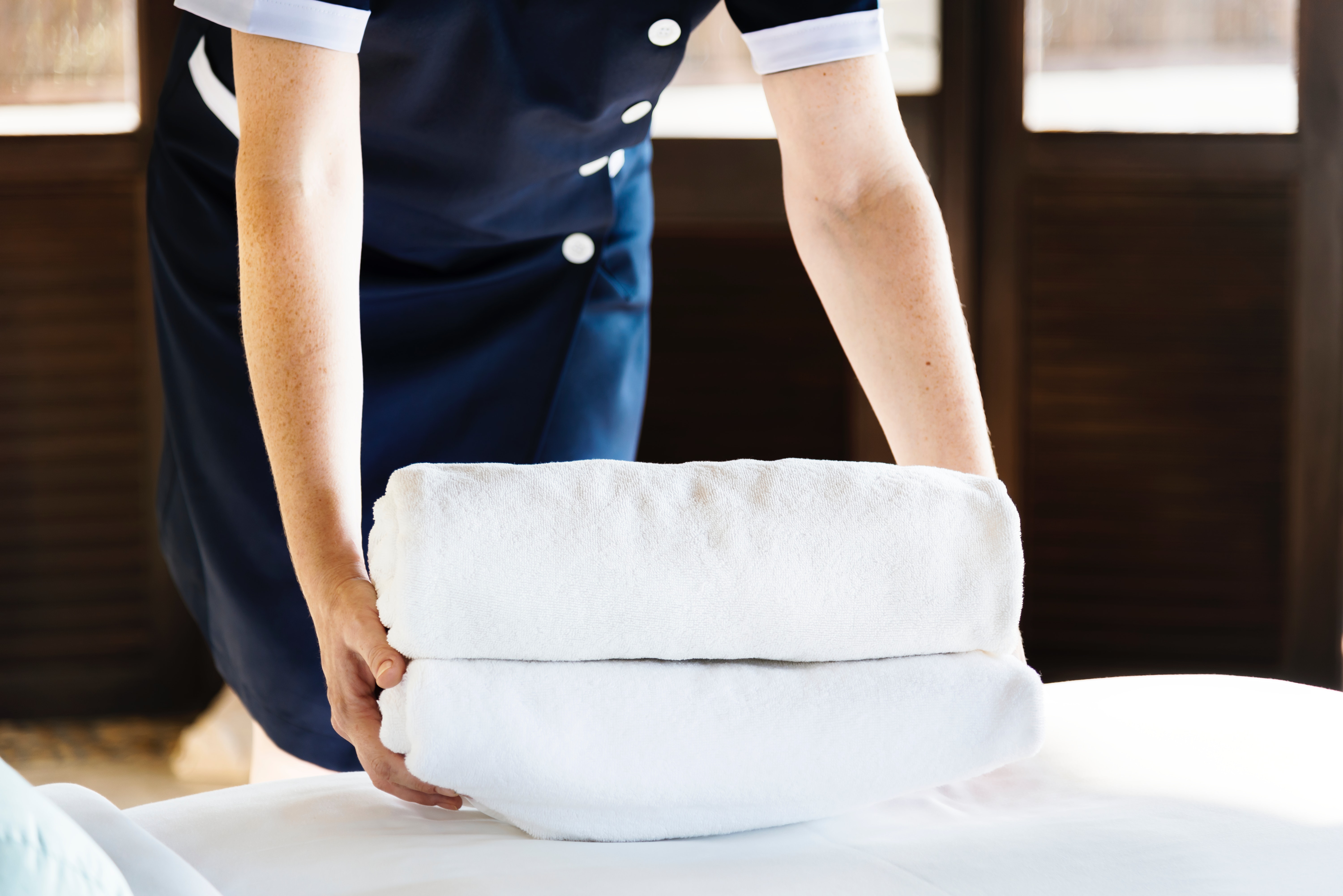A person in housekeeping uniform is folded towels on a pressed bed